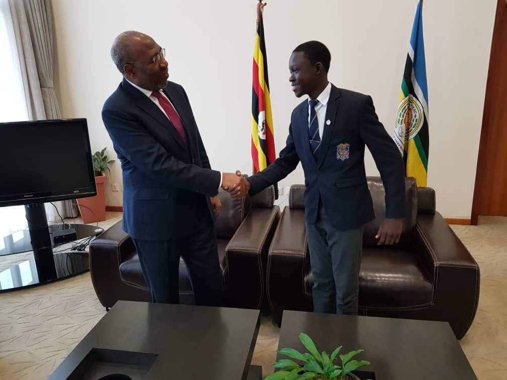 Two men shaking hands in a formal room, a Ugandan flag and an East African Community flag behind them