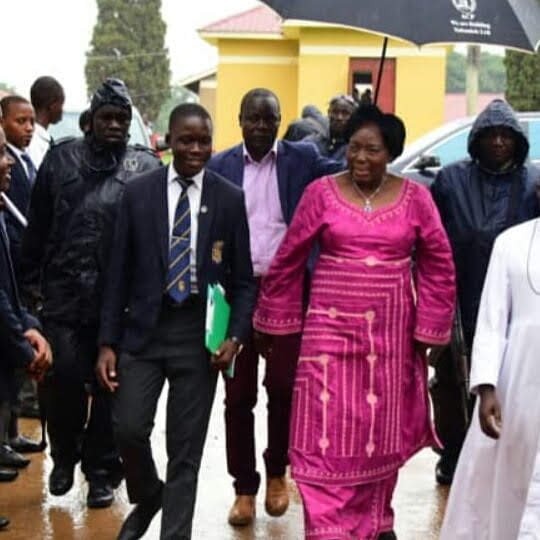 A schoolboy in blazer and tie walking with a woman in a magenta gown under an umbrella on a rainy day