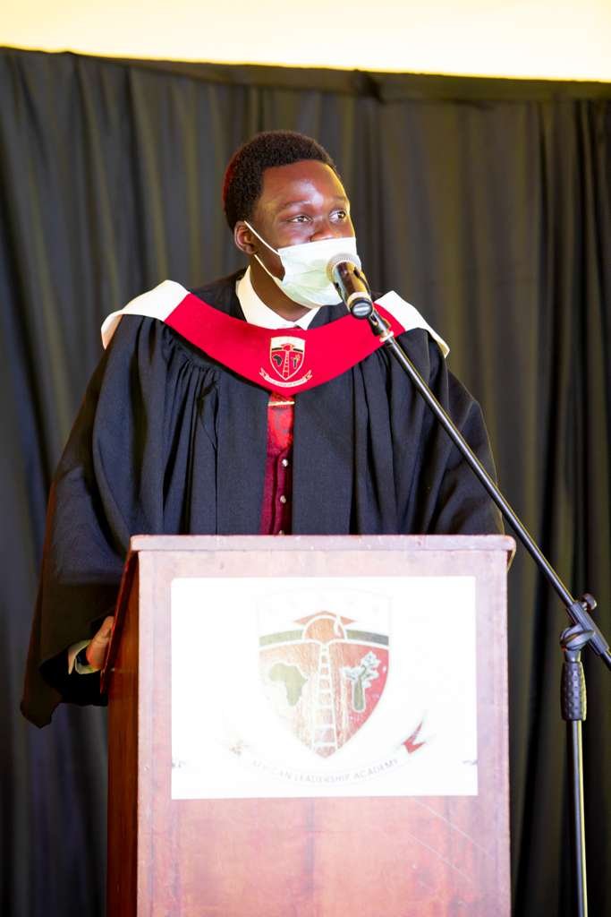 Olemo Gordon Brian in academic regalia and a face mask, speaking into a microphone from a lectern
