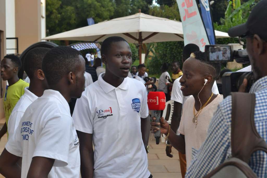 A young man in a white polo shirt being interviewed outdoors, speaking into a microphone held by a reporter